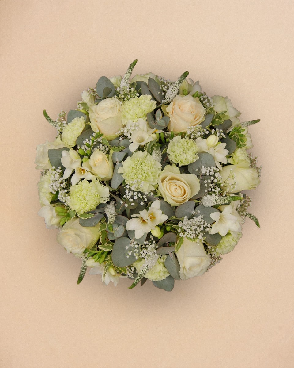 Livraison fleur deuil : Bouquet circulaire de fleurs blanches composé de roses, d œillets et de petites fleurs de gypsophile, arrangé avec des feuilles vertes, sur fond beige uni.
