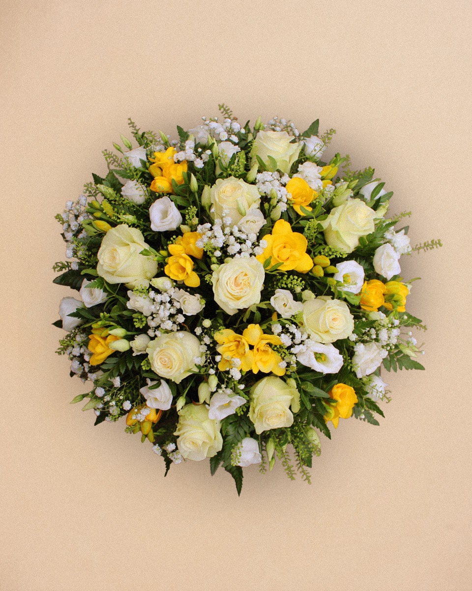Un bouquet circulaire de roses blanches, de freesias jaunes et de petites fleurs blanches. Il repose sur un fond beige.