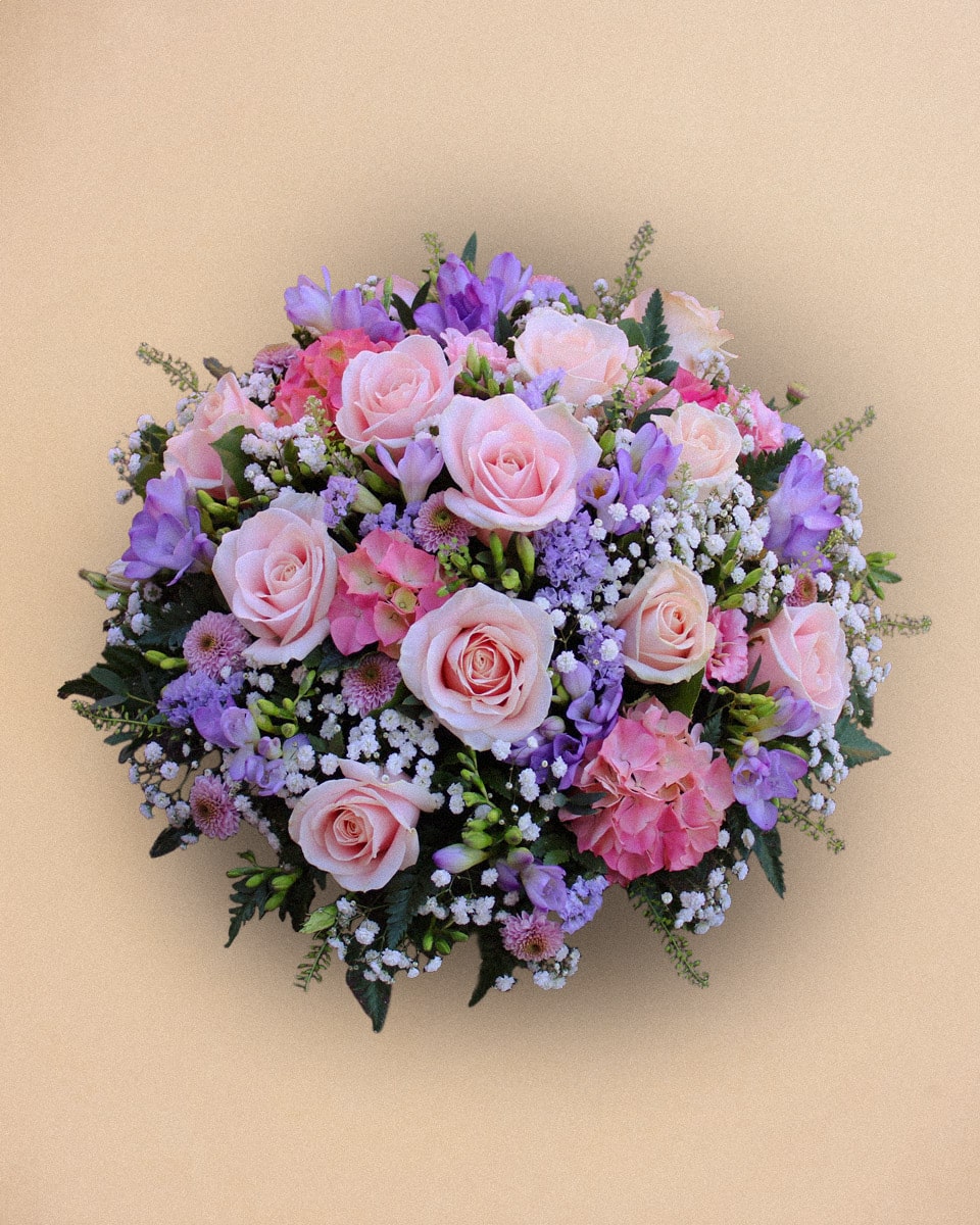 Un bouquet rond de roses roses, de freesias violets et de petites fleurs blanches est disposé sur un fond beige.